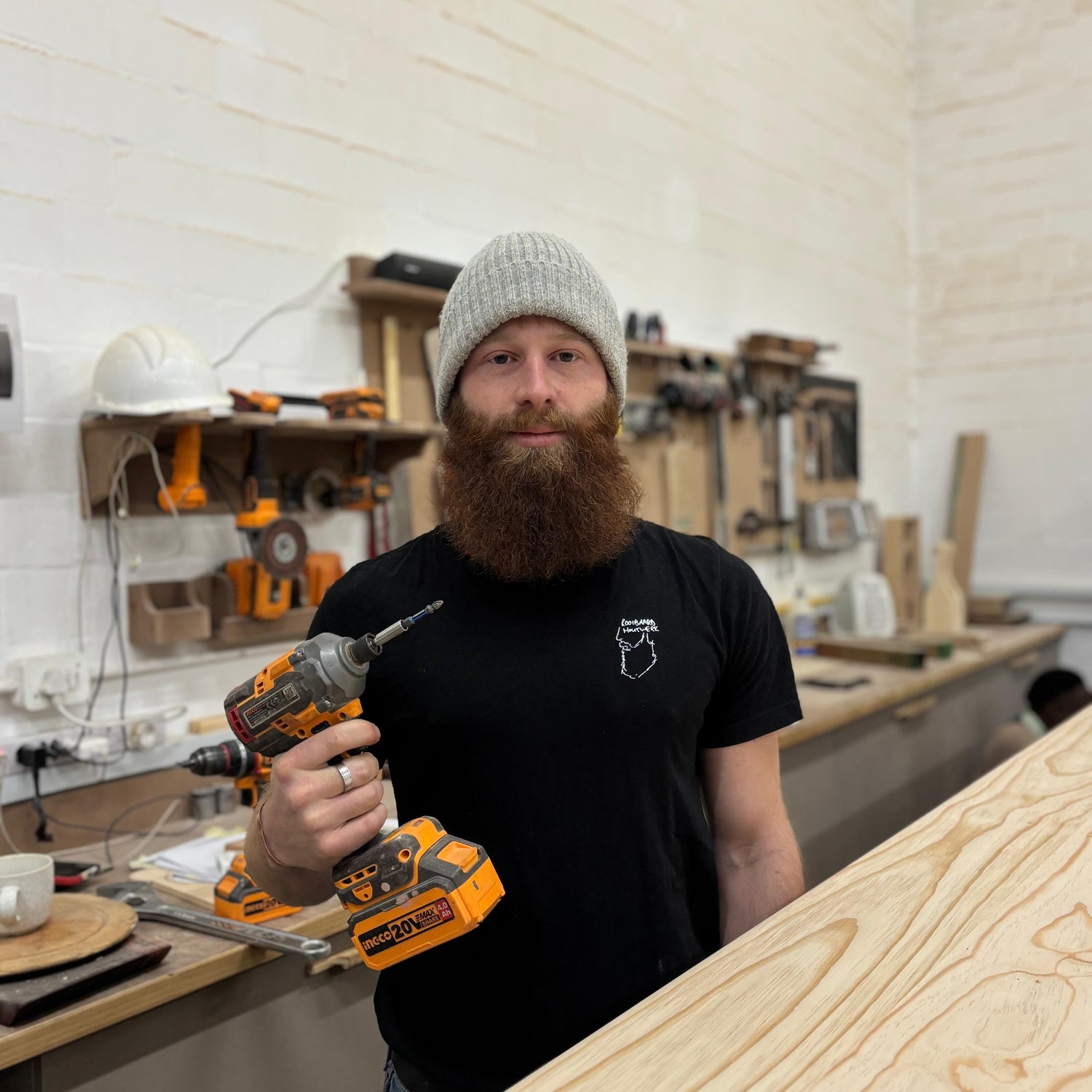 A bearded man wearing a gray knit beanie and a black t-shirt stands in a woodworking shop holding a cordless drill. The background shows a white brick wall and workbenches with tools.